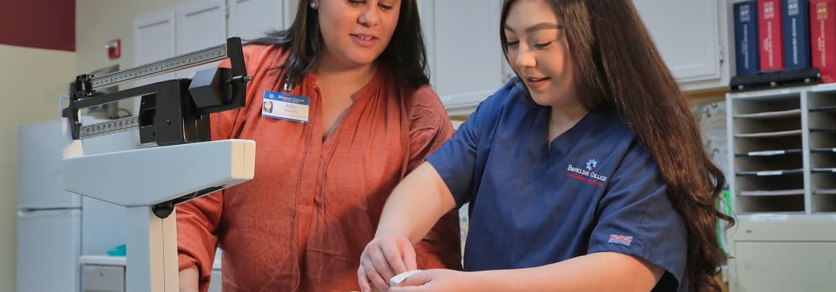 Student practicing measuring a baby's head with a baby simulation mannequin. Instructor looking on.