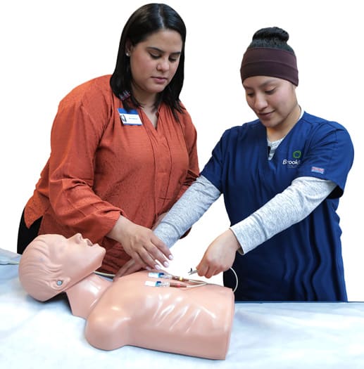 ARTY3223_logo_crop_sillo Student practicing nursing skills on a mannequin with the help of an instructor.